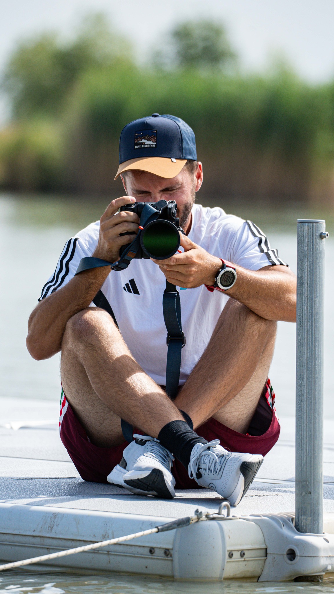 Man sitter på en EasyFloat brygga med benen i kors bredvid en stolpe och fotograferar mot kameran, på lugnt vatten.