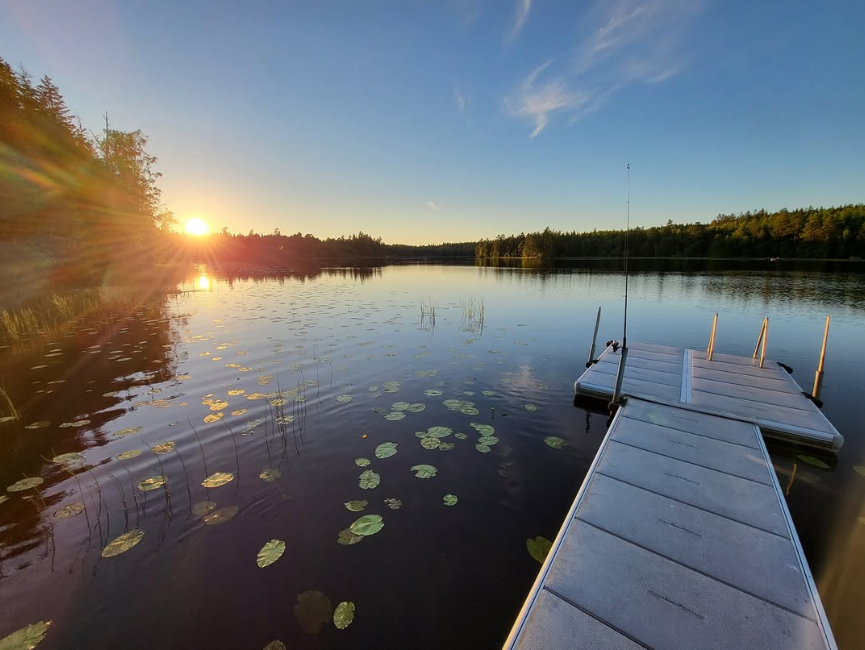 Privat EasyFloat-brygga med badstege på stilla vatten vid solnedgång. Näckrosor flyter nära bryggan och ett fiskespö lutar mot bryggans kant.